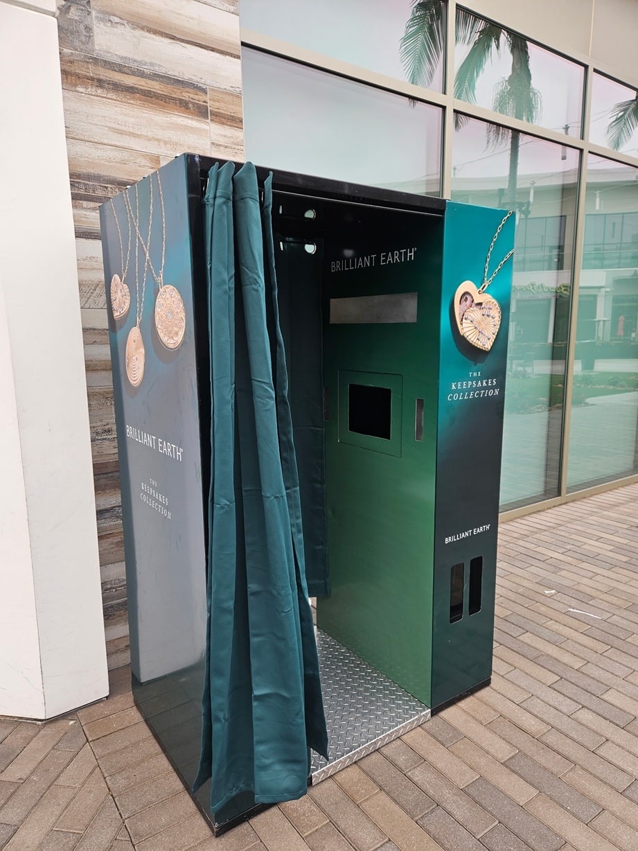 Promotional photo booth for Brilliant Earth Keepsakes Collection, teal curtains drawn open beside a storefront on a brick-paved walkway.