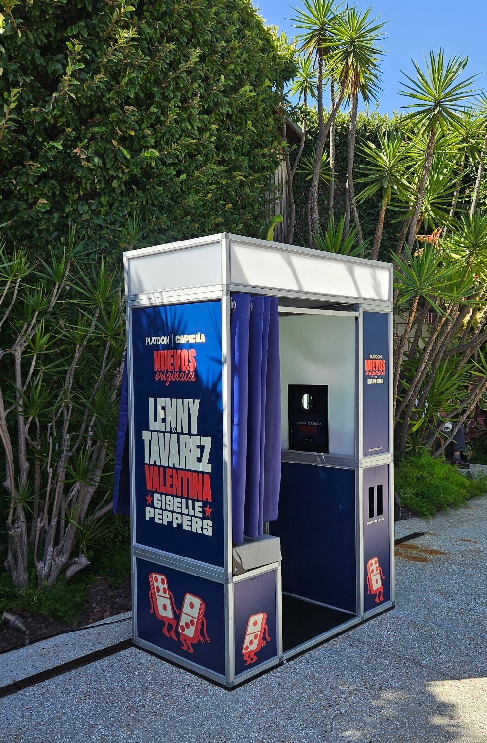Portable photo booth with blue panels displaying artist names (Lenny Tavarez, Valentina, Giselle Peppers) set outdoors among tropical plants.