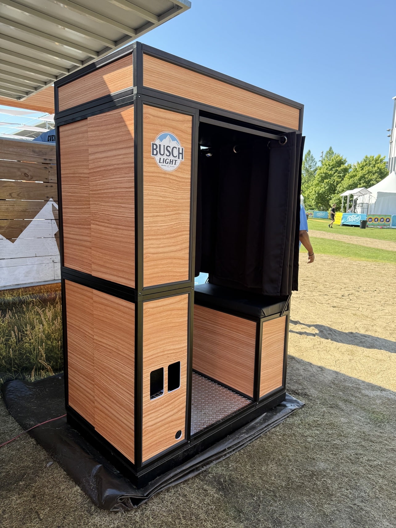 Wood-paneled photo booth with a black metal frame, open curtains, set up at an outdoor event under a clear blue sky.
