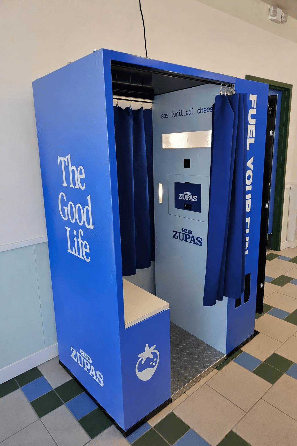 Blue photo booth capsule labeled 'The Good Life' with blue curtains and Zupas cafe branding inside a tiled indoor hallway.