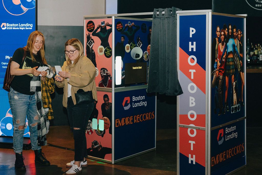 Two women stand at a photo booth area, smiling and looking at photos, with colorful posters and 'PHOTOBOOTH' signage nearby.